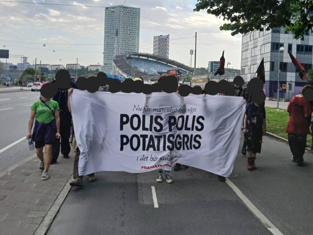 Foto av demonstrationen med en stor frontbanderoll med texten "nu får man visst inte säga POLIS POLIS POTATISGRIS i det här jävla landet..."